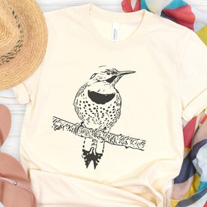 May include: A white t-shirt with a black and white illustration of a bird perched on a branch. The bird has a black head and a spotted body. The shirt is laid out on a white wooden surface with a straw hat, flip flops, and a colorful scarf.