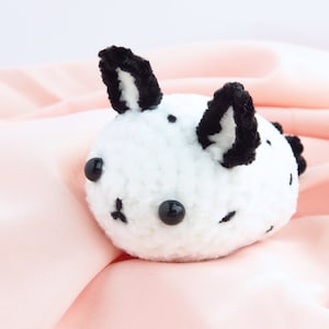 May include: White crocheted plush toy with black yarn ears and black button eyes. The toy is shaped like a small animal with a round body and a short snout.