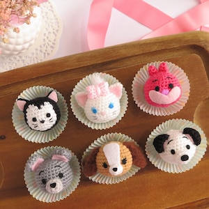 Puede incluir: Seis cabezas de personajes de crochet en moldes para cupcakes en una bandeja de madera. Los personajes son un gato blanco y negro, un gato blanco con un lazo rosa, un conejito rosa, un perro marrón y blanco, un perro blanco y negro y un ratón gris.
