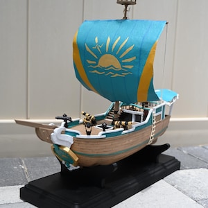 May include: A detailed model ship with a blue and gold sail featuring a sun design. The ship has a wooden hull, cannons, and a black base. The ship's dimensions are approximately 12 inches long, 4 inches wide, and 10 inches tall.