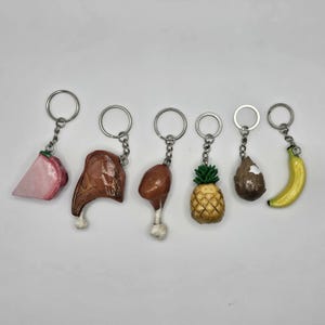 May include: Six different food-shaped keychains. The keychains are made of plastic and feature a pineapple, banana, chicken leg, ham, coconut, and a piece of ham. The keychains are all attached to silver keyrings.