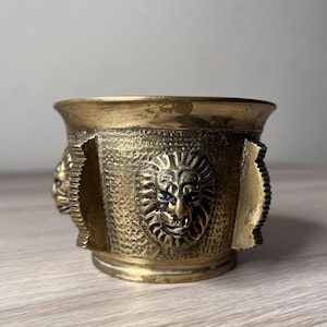 May include: A brass mortar featuring a textured surface and three-dimensional lion head motifs positioned around the exterior. The vessel has a wide, flared rim and a cylindrical body with vertical ridges separating each lion head detail.