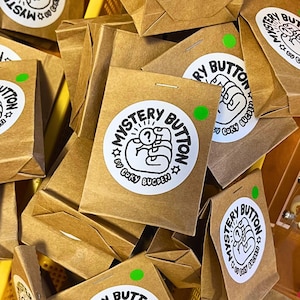 May include: A pile of small, brown paper bags, each sealed with a staple and a round sticker. The stickers read "Mystery Button" with a hand holding a question mark. A small green dot is on each bag.