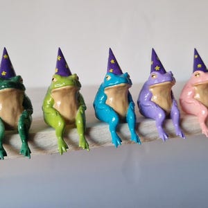May include: Five frog figurines wearing purple wizard hats with yellow stars. The frogs are sitting on a wooden surface. The frogs are green, blue, purple, and pink.