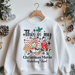 May include: White sweatshirt featuring a festive Christmas graphic. The design includes gingerbread men, Christmas trees, a snowman in a mug, and the text "This is my Christmas Movie Watching Shirt."