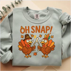 May include: A light blue sweatshirt with the text "OH SNAP!" in orange. The graphic features two cartoon turkeys holding a wishbone, with fall leaves. One turkey wears a pilgrim hat, the other a feather headdress.