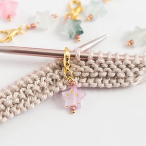 Könnte beinhalten: Ein rosafarbener sternförmiger Maschenmarkierer mit goldenen Flocken auf einem beigefarbenen Strickstoff. Der Markierer ist an einem goldenen Ring und einer Stricknadel befestigt.