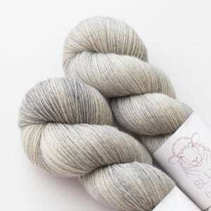 May include: A skein of soft, light gray and white variegated yarn with a subtle heathered texture. The yarn is wrapped with a white paper label featuring a line-art illustration of a sheep and the partial text BLU.