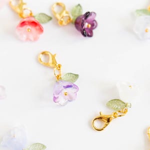 Pode incluir: Conjunto de pingentes de flores coloridas com fechos dourados. Os pingentes apresentam flores de diferentes cores com folhas verdes.