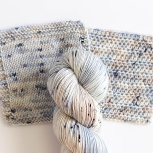 May include: A ball of speckled yarn in white, blue, and tan, beside two knitted swatches. The yarn has a soft, textured appearance, and the swatches show the knitted stitch pattern. The background is white.