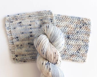 May include: A skein of speckled yarn in shades of white, blue, and tan, next to two knitted swatches. The yarn has a soft, textured appearance, and the swatches show the knitted stitch pattern. The background is white.