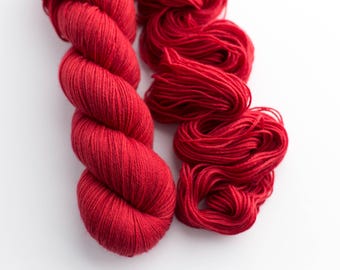 May include: Two skeins of vibrant red yarn are displayed against a white background. One skein is tightly wound, while the other is loosely arranged. The yarn's texture is visible, suggesting a soft, high-quality material. Ideal for knitting or crochet projects.