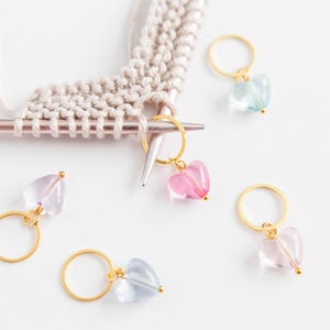 May include: A collection of heart-shaped stitch markers in various colors, including pink, blue, and purple, with gold-colored rings. The markers are placed near a knitting needle with a partially completed knitted piece.