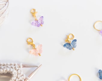 May include: Five gold butterfly stitch markers with clear butterfly charms in pink, purple, and blue. The markers are on a white surface.