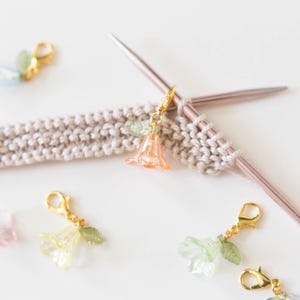 May include: A set of five flower-shaped stitch markers for knitting or crocheting. The markers are made of clear plastic with gold-toned metal clasps and feature different colored flowers.