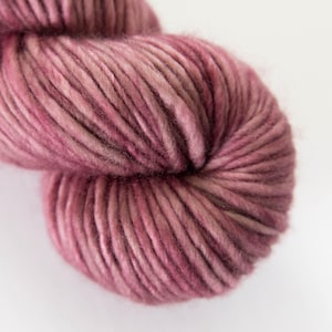 May include: Close-up of a skein of chunky yarn in shades of dusty rose and mauve. The yarn is thick and textured, with visible fibers. The background is plain white, highlighting the yarn's color and texture. Ideal for knitting and crochet projects.