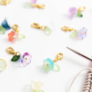 May include: A collection of colorful flower-shaped stitch markers with gold-colored clasps and green leaf accents. The markers are in shades of red, blue, purple, and yellow. Knitting needles and yarn are also visible.