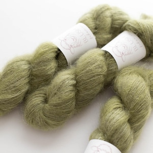 May include: Close-up of several skeins of olive green yarn. The yarn is soft and fluffy, with a slight sheen. Each skein is wrapped with a white label featuring a floral design and the text "BLUE SKY FIBERS".