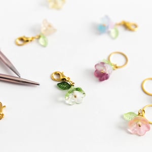 Könnte beinhalten: Sortiment an bunten, blumenförmigen Maschenmarkierern mit goldenen Ringen und Verschlüssen. Die Markierer sind aus Glas und haben ein zartes, feminines Design.