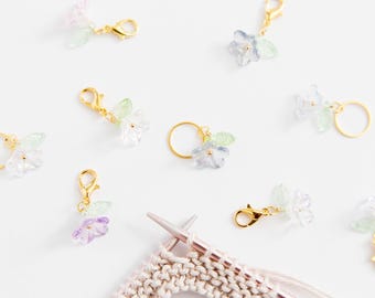 May include: A set of flower-shaped stitch markers for knitting or crocheting. The markers are made of clear plastic with a gold metal clasp and come in a variety of colors, including blue, green, pink, and purple.