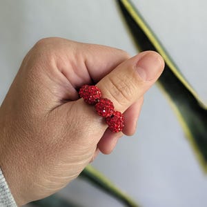May include: A red beaded ring with three round, sparkling beads. The ring is held up against a light background with a green and yellow plant in the background. The ring is a vibrant shade of red.