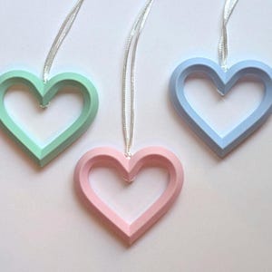 May include: Three heart-shaped ornaments in pastel colors: mint green, pink, and light blue. Each heart is suspended by a silver ribbon. The ornaments are likely for decoration, possibly for Valentine's Day or other celebrations.