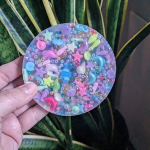 May include: A clear round resin coaster with a variety of colorful sea-themed charms embedded inside, including seashells, starfish, fish, and beads.