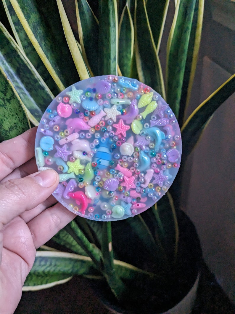 May include: A clear round resin coaster with a variety of colorful sea-themed charms embedded inside, including seashells, starfish, fish, and beads.