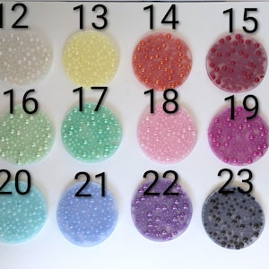 May include: A collection of twelve circular, translucent embellishments, each filled with small, colorful beads. The colors include white, yellow, orange, red, pink, green, teal, blue, purple, and black. Numbers 12 through 23 are printed above each circle.