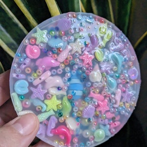 May include: A clear round resin coaster with a variety of colorful sea-themed charms embedded inside, including seashells, starfish, fish, and seahorses.