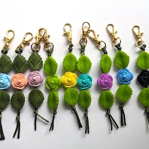 May include: A collection of keychains with gold-colored clasps. Each keychain features a green leaf design, a colorful flower, and a green tassel. The flowers are in various colors, including blue, pink, yellow, and white.