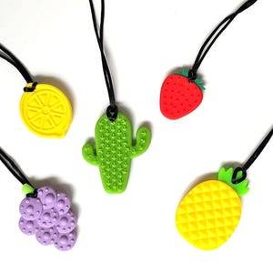 May include: A collection of five colorful silicone necklaces with fruit and plant-themed pendants. The pendants include a yellow lemon, a green cactus, a red strawberry, a purple grape cluster, and a yellow pineapple. Each pendant hangs from a black cord.