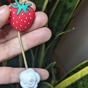 Op de afbeelding: Een rode en groene aardbeienbedel met een witte rozenbedel bevestigd aan een beige koord.