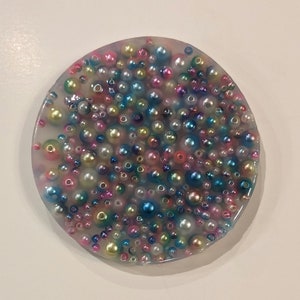 May include: A round, clear resin coaster filled with a multitude of colorful beads. The beads vary in size and color, including pink, blue, green, and gold, creating a textured, decorative surface. The coaster is a unique, handcrafted item.
