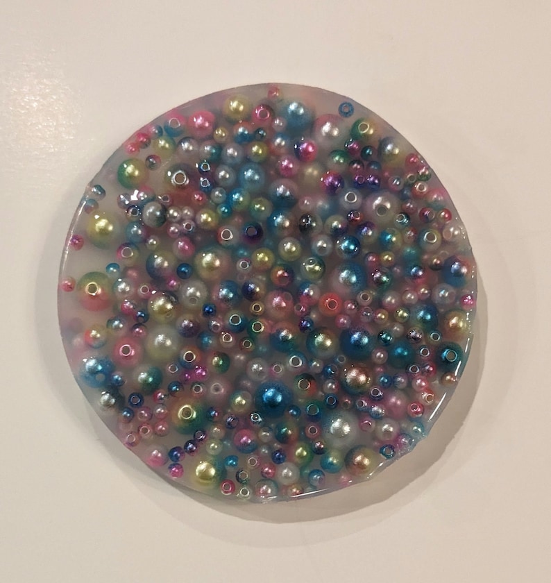 May include: A round, clear resin coaster filled with a multitude of colorful beads. The beads vary in size and color, including pink, blue, green, and gold, creating a textured, decorative surface. The coaster is a unique, handcrafted item.