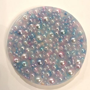 May include: A round, clear resin coaster filled with various sizes of iridescent pearls. The pearls are in shades of white, pink, blue, and silver, creating a shimmering effect. The coaster has a smooth, glossy finish.