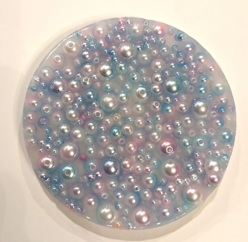 May include: A round, clear resin coaster filled with various sizes of iridescent pearls. The pearls are in shades of white, pink, blue, and silver, creating a shimmering effect. The coaster has a smooth, glossy finish.