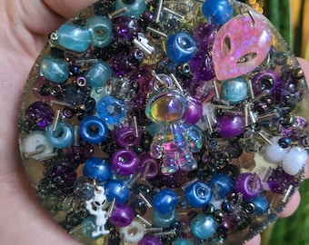 May include: A round, clear resin coaster filled with colorful beads and charms. The coaster features blue, purple, and clear beads, along with small alien-themed charms. A pink alien head charm and a small astronaut charm are also visible.