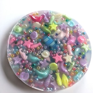 May include: A round, clear resin coaster with a variety of colorful sea-themed charms embedded inside. The charms include starfish, seashells, fish, and pearls.