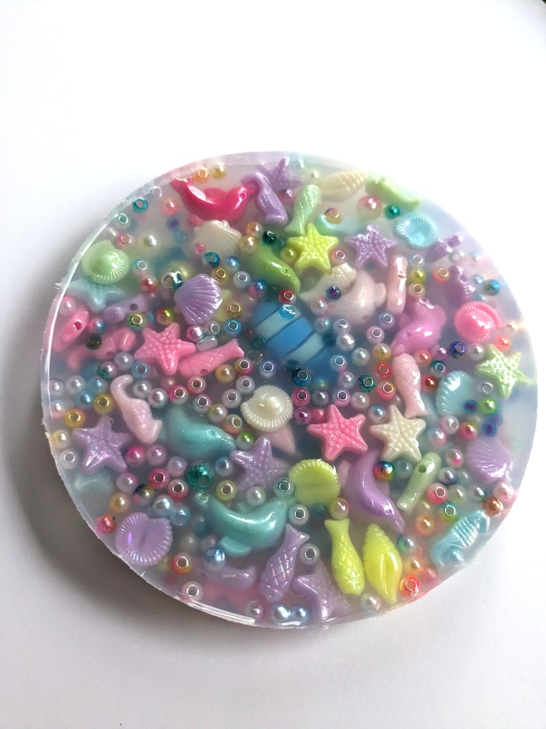 May include: A round, clear resin coaster with a variety of colorful sea-themed charms embedded inside. The charms include starfish, seashells, fish, and pearls.