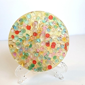May include: A round, gold-colored coaster with embedded colorful sequins. The sequins are in shades of red, green, yellow, pink, and blue. The coaster is displayed on a clear plastic stand, suitable for home decor.