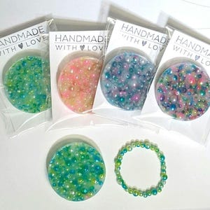 May include: A collection of handmade resin coasters in various colors, each embedded with small, colorful beads. The coasters are packaged with a white card that reads "HANDMADE WITH LOVE". A matching beaded bracelet is also included.