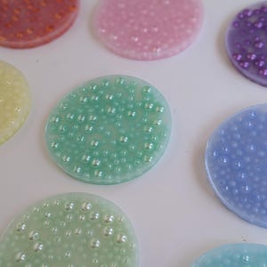 May include: A collection of colorful, oval-shaped resin coasters. The coasters are in various pastel colors, including pink, green, blue, and yellow. Each coaster has a textured surface with small, embedded, pearl-like beads.