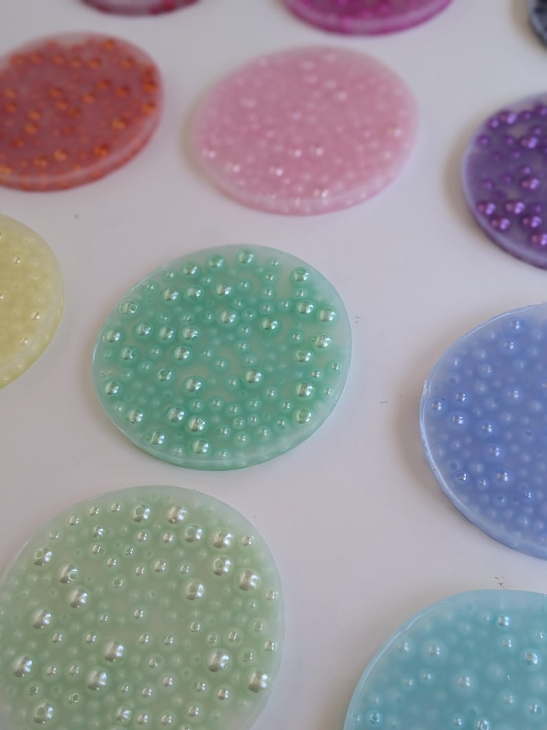 May include: A collection of colorful, oval-shaped resin coasters. The coasters are in various pastel colors, including pink, green, blue, and yellow. Each coaster has a textured surface with small, embedded, pearl-like beads.