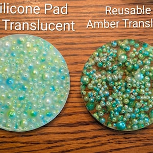 May include: Two round, translucent pads covered in small, colorful beads. One is clear with blue and green beads, the other is amber with green and yellow beads. Text at the top reads "Silicone Pad Clear Translucent" and "Reusable Pad Amber Translucent."