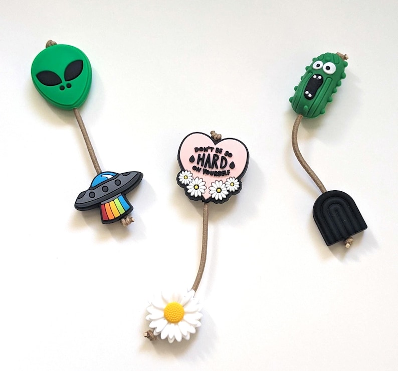 Op de afbeelding: Drie kleurrijke siliconen bedels met een touwtje aan elk. De bedels zijn een groene alien, een roze hart met de tekst "Wees niet zo hard voor jezelf" en een groene cartoon cactus.
