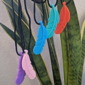 May include: A collection of feather-shaped pendants in various colors, including blue, teal, orange, pink, and purple, suspended from black cords. The pendants are displayed against a backdrop of green and yellow plant leaves.