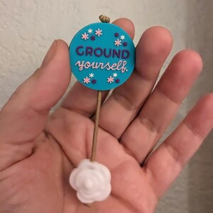 Op de afbeelding: Een ronde blauwe en witte hanger met de tekst "GROUND yourself" en roze en paarse bloemen. De hanger is bevestigd aan een witte roosvormige hanger met een bruin touwtje.
