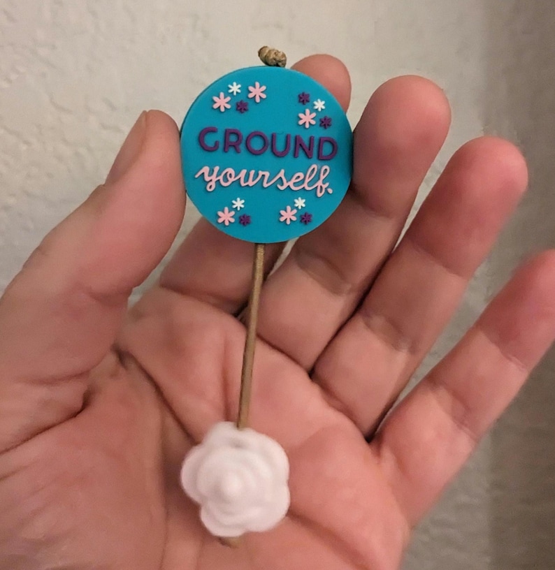 Op de afbeelding: Een ronde blauwe en witte hanger met de tekst "GROUND yourself" en roze en paarse bloemen. De hanger is bevestigd aan een witte roosvormige hanger met een bruin touwtje.