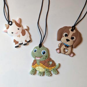 May include: Three animal-shaped pendants on black cords. The pendants include a white and brown goat, a green and brown turtle, and a brown and white puppy. Each pendant has a black cord for wearing.